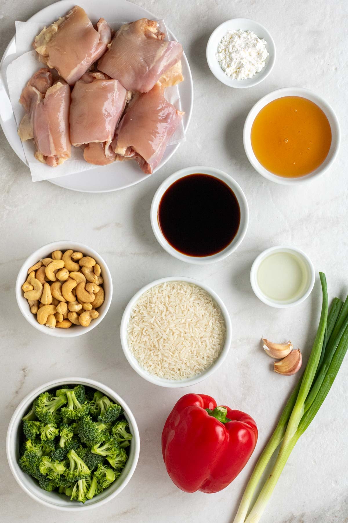 Plate of raw chicken thighs with bowls of cornstarch, honey, soy sauce, rice vinegar, white rice, roasted cashews, and broccoli florets with garlic cloves, green onions, and a red bell pepper on a white background.