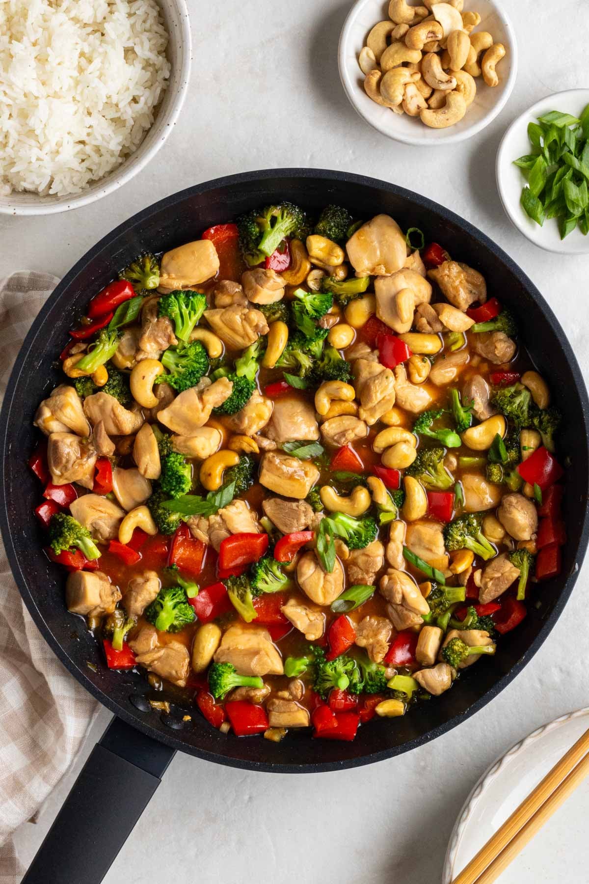 Large pan of honey cashew chicken with a bowl of cooked white rice, roasted cashews, and sliced green onions on the side with an empty bowl with wood chopsticks on a white background.