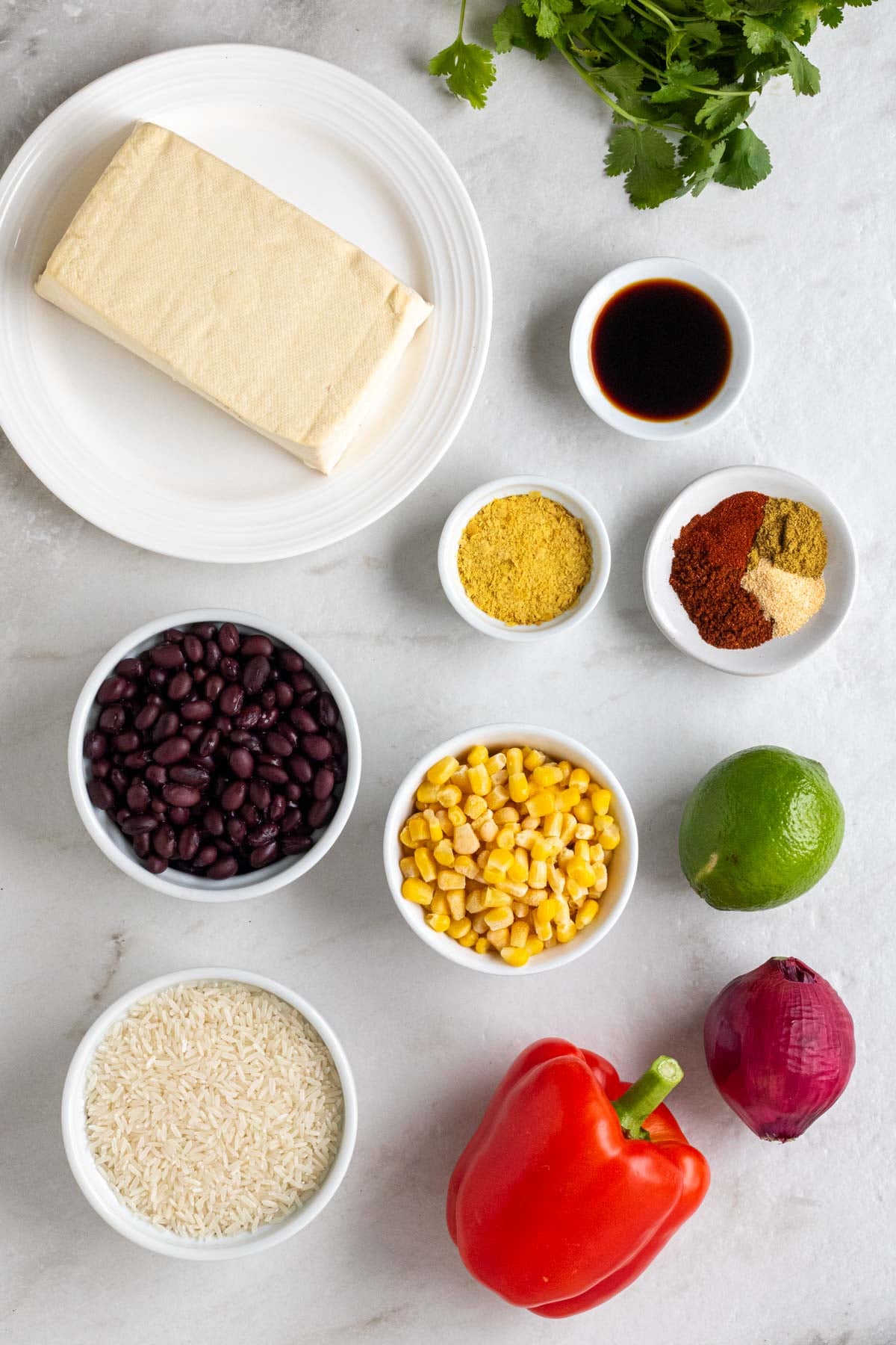 Plate with a block of tofu, fresh cilantro, bowl of soy sauce, bowl of nutritional yeast, bowl of spices, bowl of black beans, bowl of frozen corn, bowl of whit rice, with a lime, red onion, and red bell pepper on a white background.