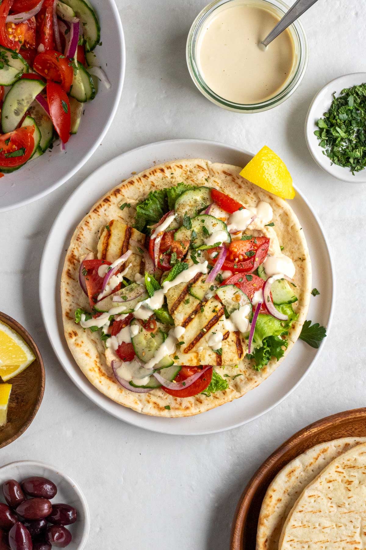 An open Mediterranean halloumi wrap with garlic sauce drizzled on top with extra garlic sauce, parsley, tomato cucumber salad, pitas, lemon wedges, and olives on the side.