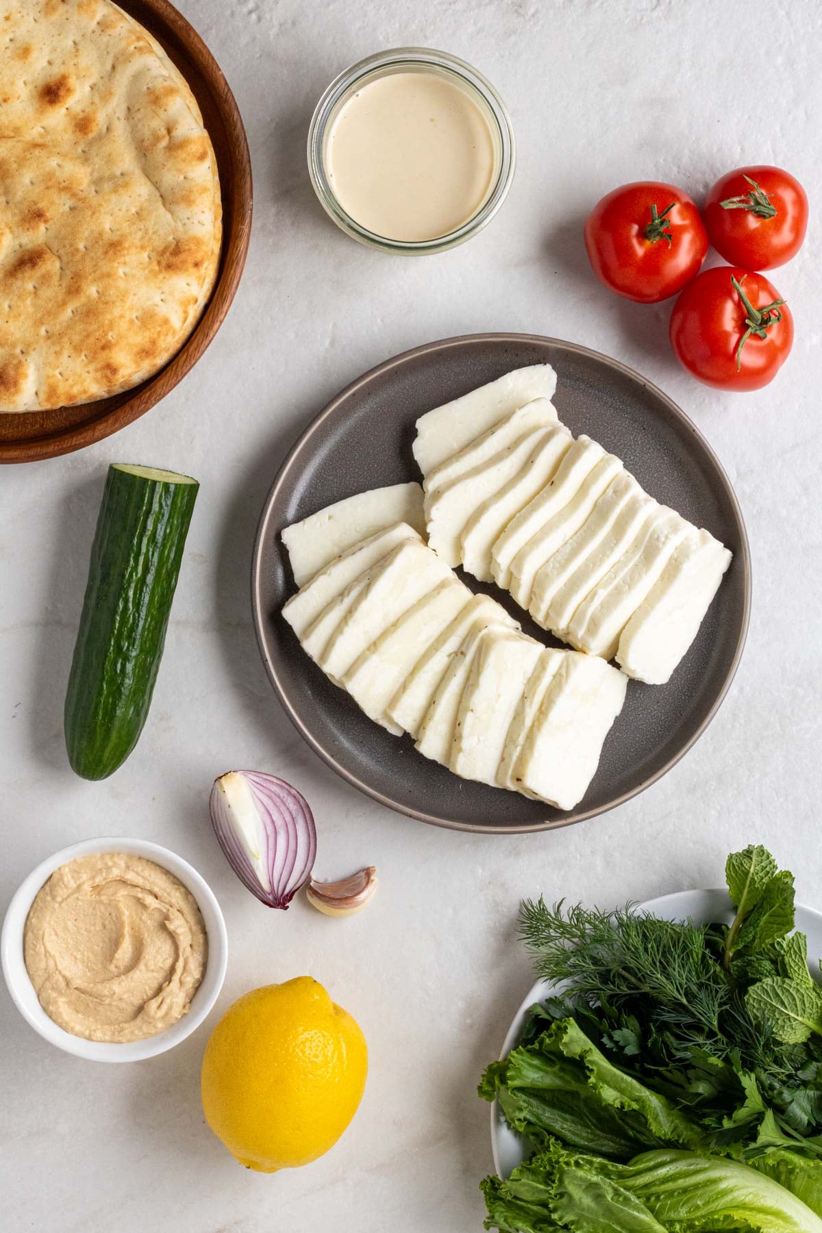 Pitas on a wood plate, a bowl of garlic sauce, ripe tomatoes, plate of sliced halloumi cheese, cucumber, red onion, garlic cloves, bowl of hummus, lemon, and bowl of fresh herbs on a white background.