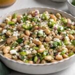 Close up of a white scalloped bowl of white bean salad with a bowl of crumbled feta cheese and jar of salad dressing in the background.