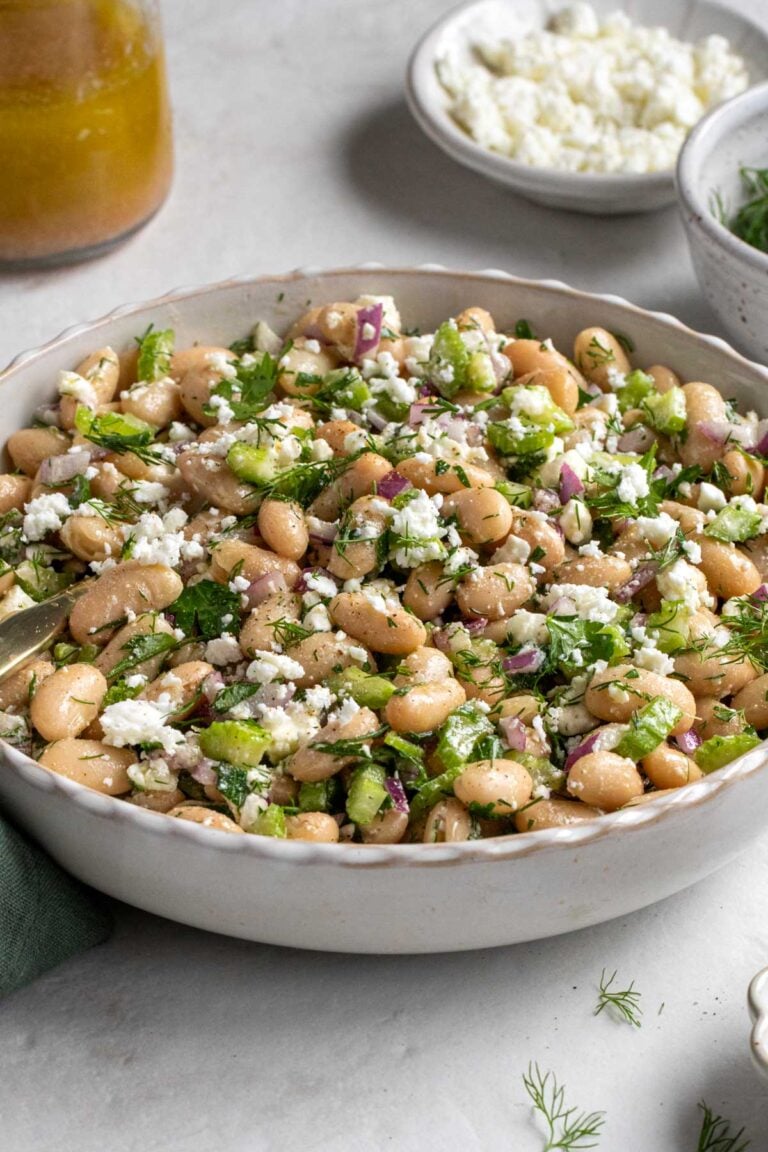 Close up of a white scalloped bowl of white bean salad with a bowl of crumbled feta cheese and jar of salad dressing in the background.
