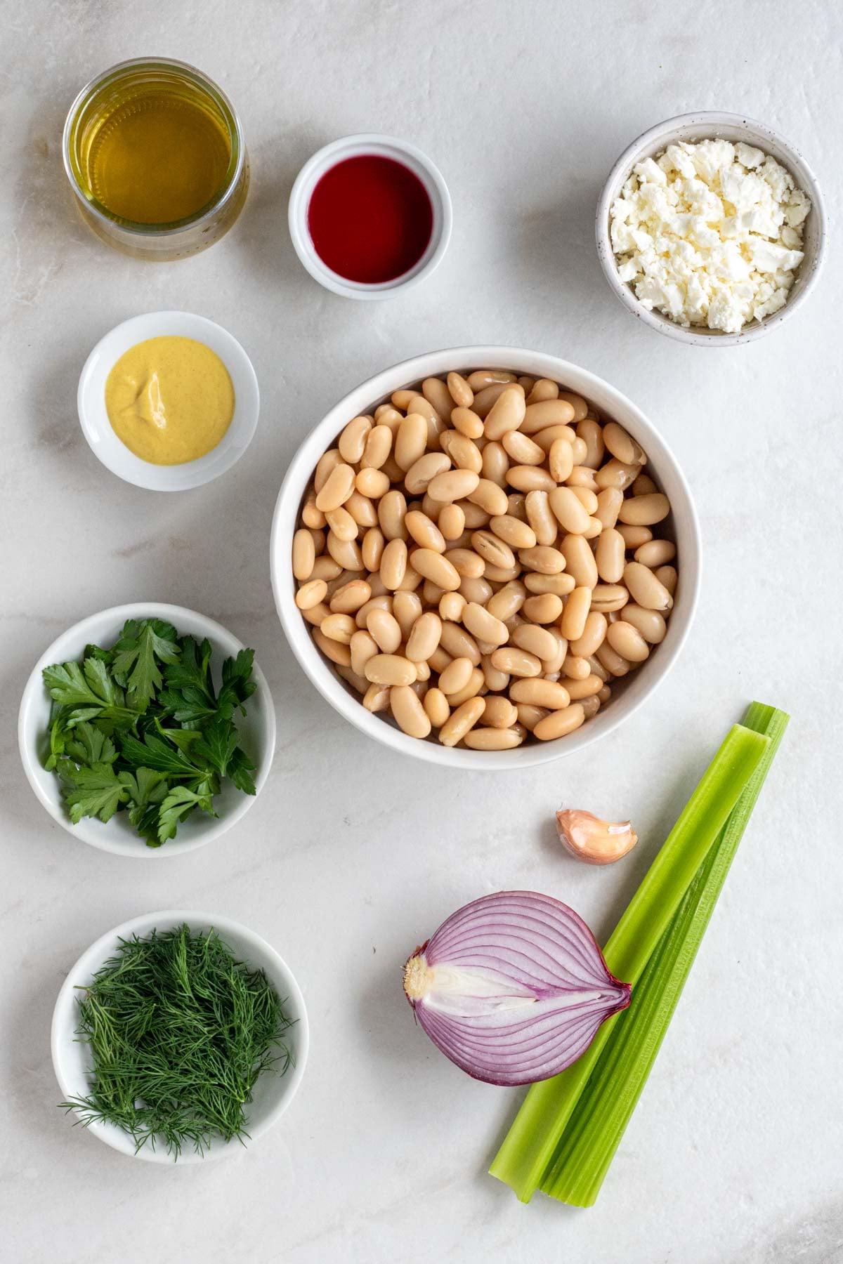 Jar of olive oil with bowls of red wine vinegar, Dijon mustard, feta cheese, canned white beans, fresh parsley, and fresh dill with a clove of garlic, half a red onion, and two celery stalks on a white background.