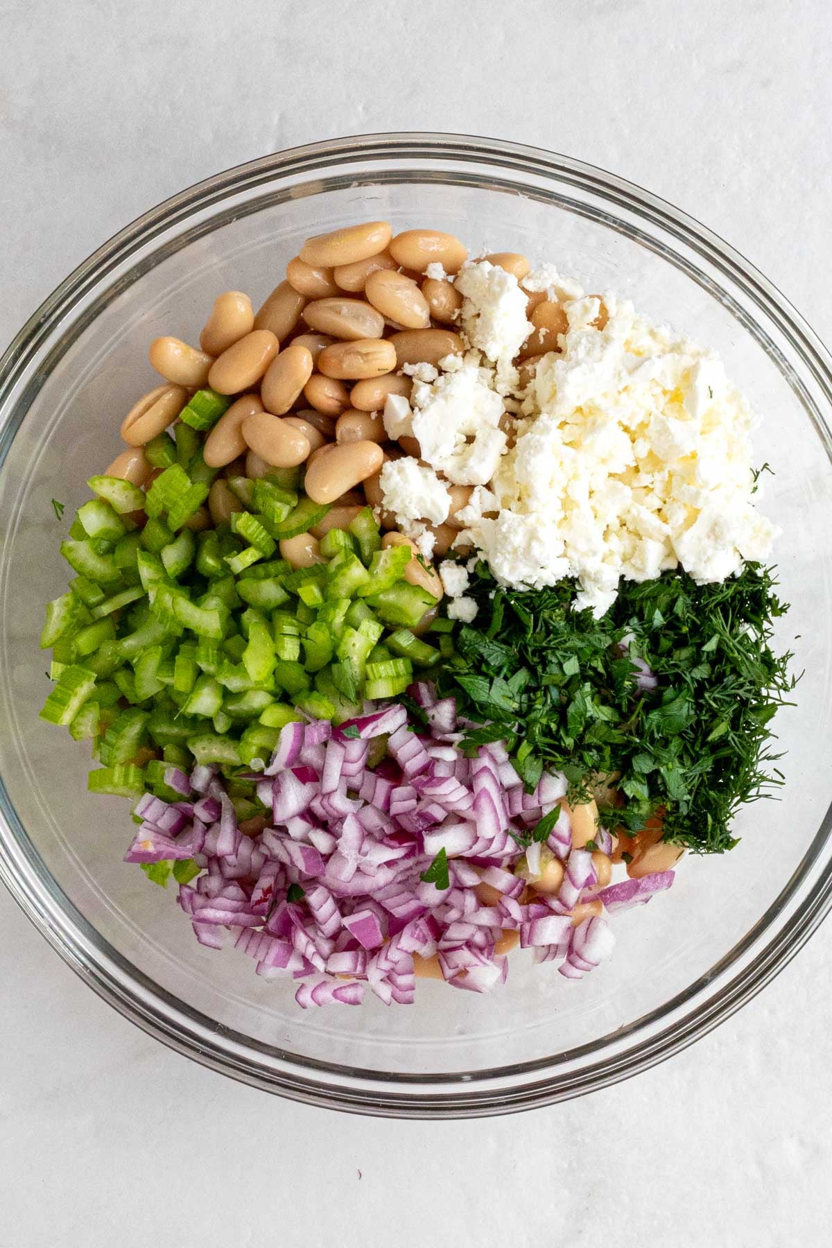 Glass bowl of white beans, diced celery, diced red onion, chopped herbs, and crumbled feta cheese.