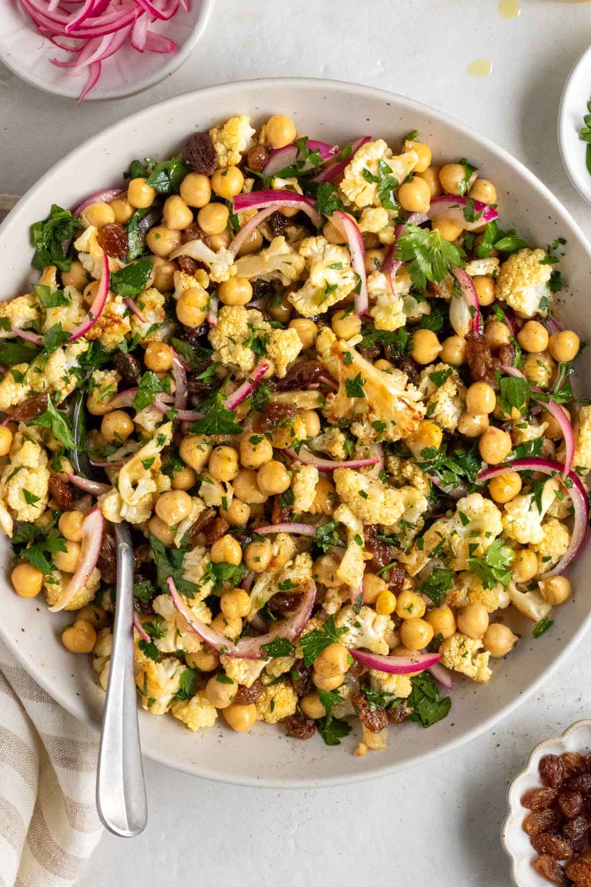 Large bowl of roasted cauliflower and chickpea salad with a serving spoon with small bowls of red pickled onions, fresh parsley, and raisins on the side.
