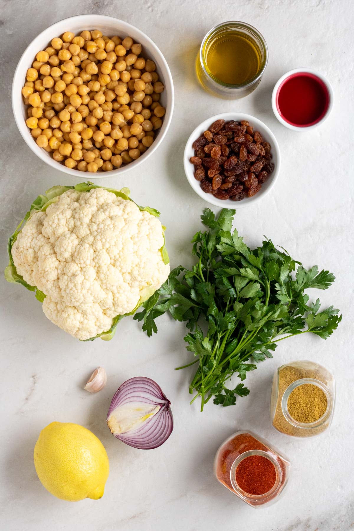 Large bowl of chickpeas, jar of olive oil, bowl of red wine vinegar, bowl of raisins, head of cauliflower, fresh parsley, red onion, garlic, lemon, jar of cumin, and jar of smoked paprika on a white background.