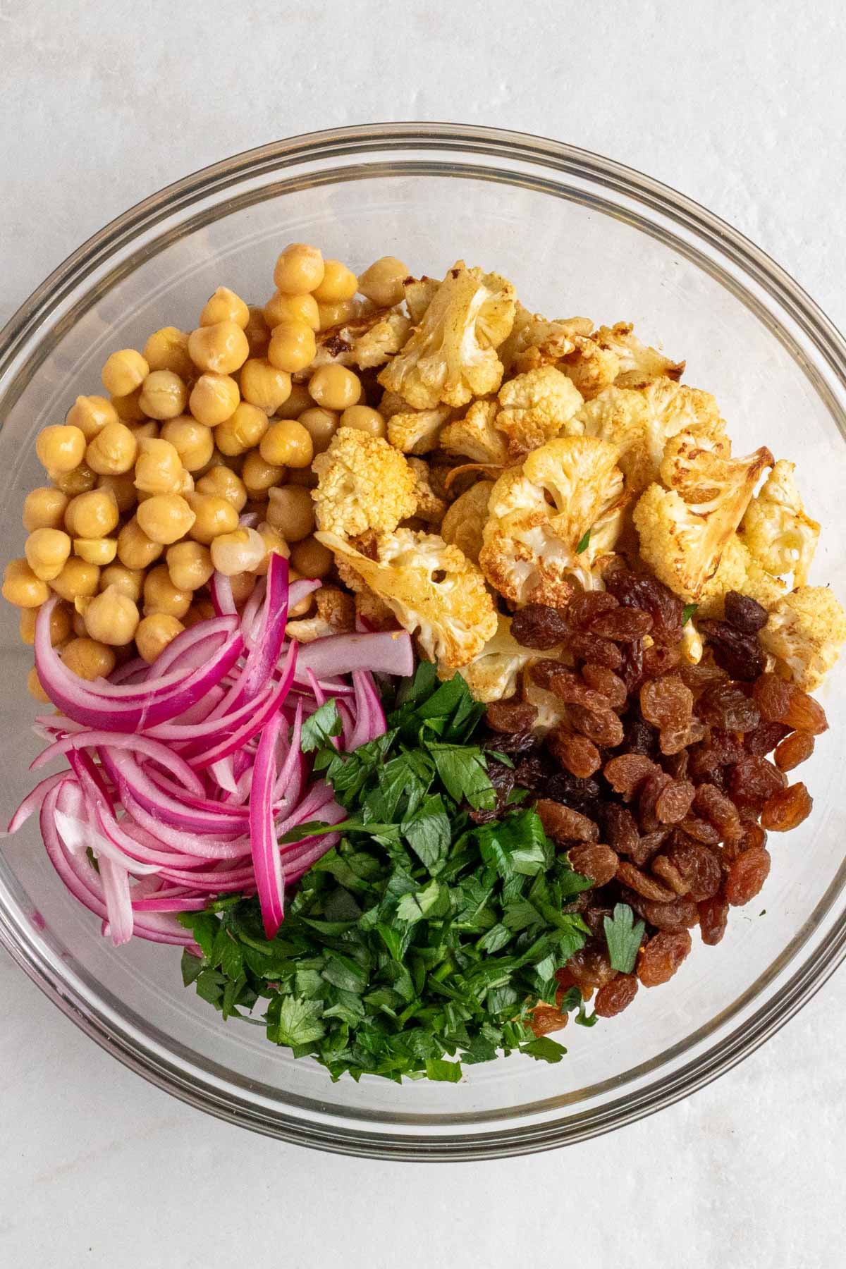 Large glass bowl filled with roasted cauliflower, chickpeas, pickled red onions, chopped parsley, and raisins on a white background.