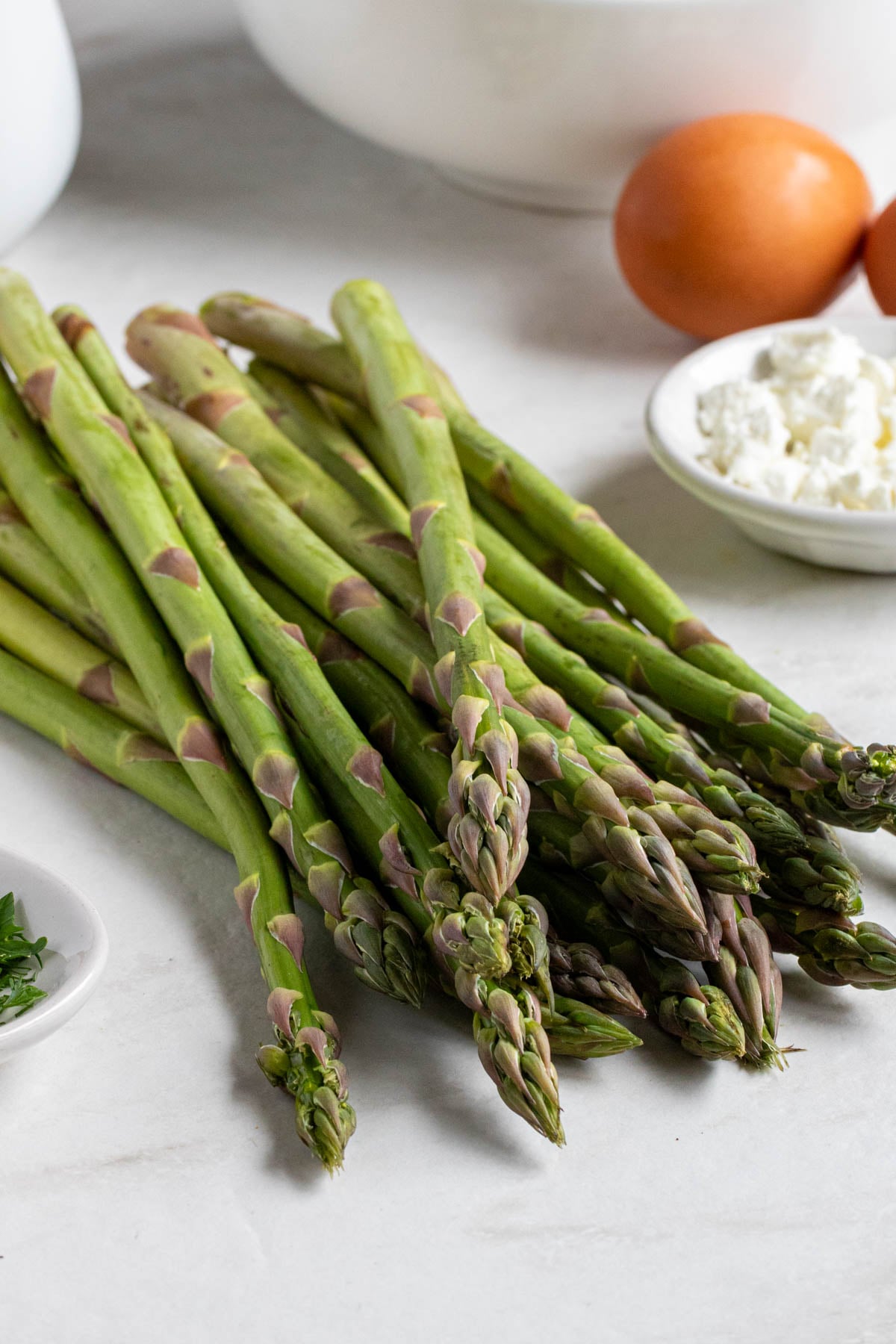 Bowls of eggs, fresh herbs, and crumbled feta cheese with a bunch of fresh, raw asparagus, an onion, and a jug of milk on a white background.