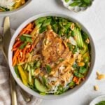 Two chicken spring roll bowls with bowls of peanut sauce, green onions, and fresh herbs on the side with gold cutlery and a white and beige checkered tea towel on the side.