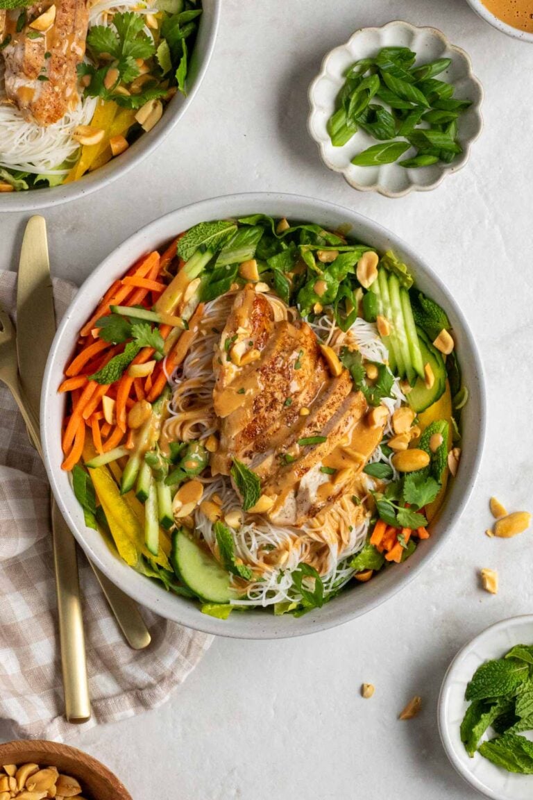Two chicken spring roll bowls with bowls of peanut sauce, green onions, and fresh herbs on the side with gold cutlery and a white and beige checkered tea towel on the side.