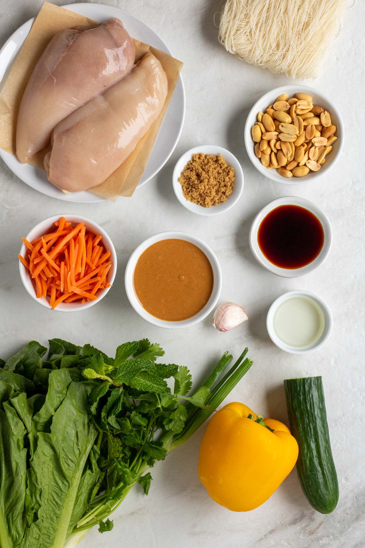 Plate of chicken breasts, vermicelli noodles, with bowls of peanuts, brown sugar, soy sauce, peanut butter, rice vinegar, and shredded carrots with garlic cloves, lettuce, fresh herbs, bell pepper, and cucumber on a white background.