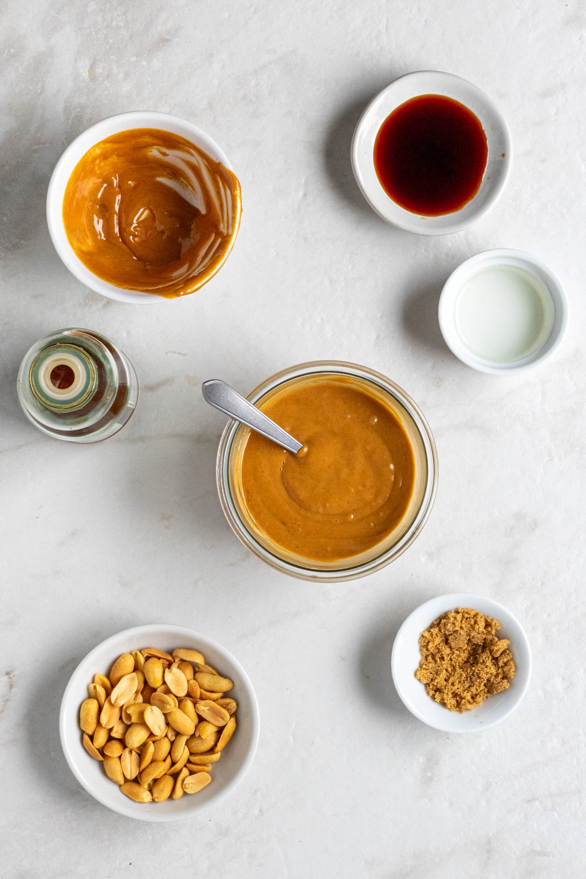A small bowl of peanut sauce with a bottle of sesame oil and bowls of creamy peanut butter, soy sauce, rice vinegar, brown sugar, and peanuts on the side on a white background.