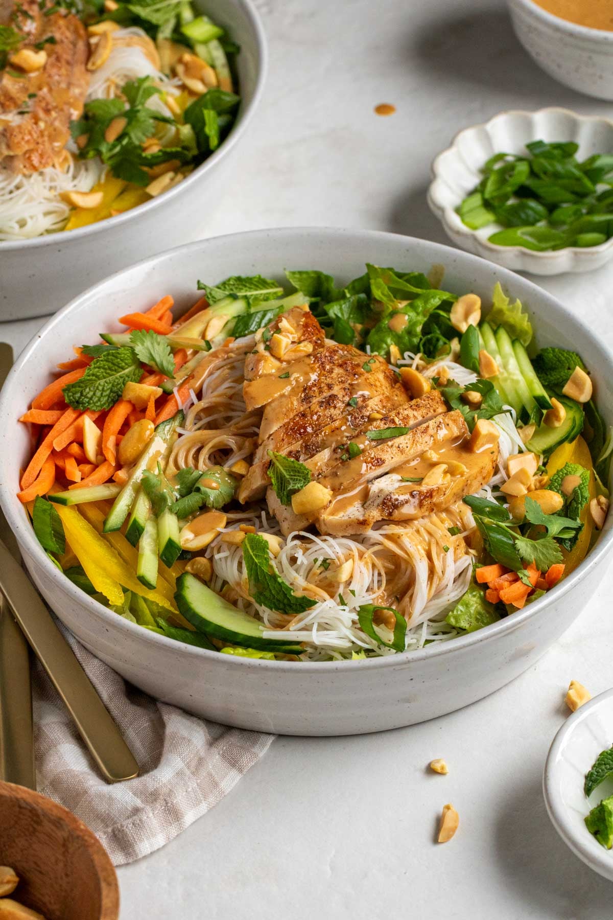 Two spring roll bowls with grilled chicken with peanut sauce and chopped peanuts on top with cutlery and a tea towel on the side.