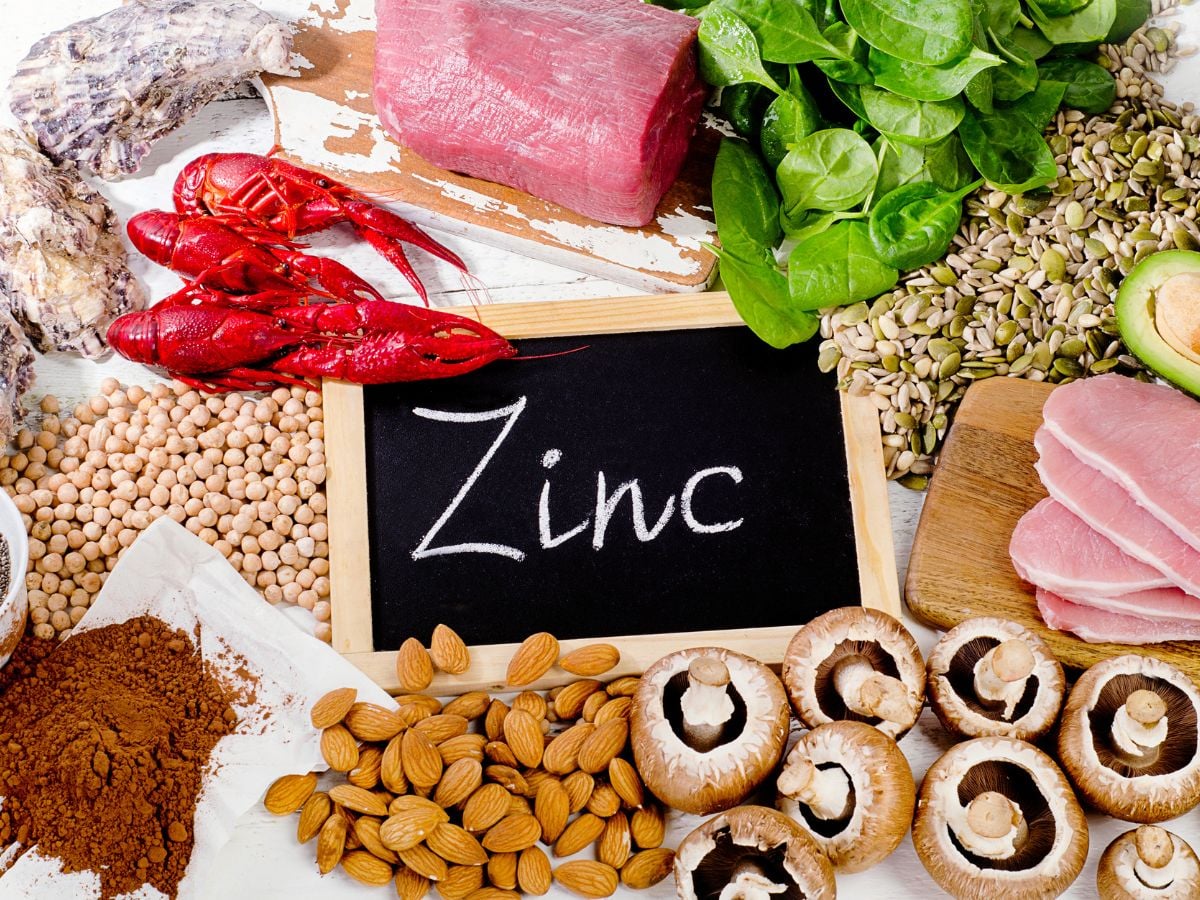 Small chalkboard with the word zinc written on it surrounded by oysters, pork, shellfish, spinach, seeds, avocado, pork, mushrooms, almonds, cocoa powder, and dried chickpeas.