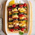 Grilled halloumi and vegetables skewers on a plate with parchment paper on a wood cutting board with a bowl of couscous, parsley, olive oil, and a white and grey striped tea towel on the side.