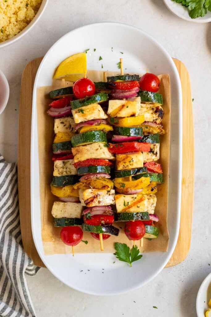 Grilled halloumi and vegetables skewers on a plate with parchment paper on a wood cutting board with a bowl of couscous, parsley, olive oil, and a white and grey striped tea towel on the side.