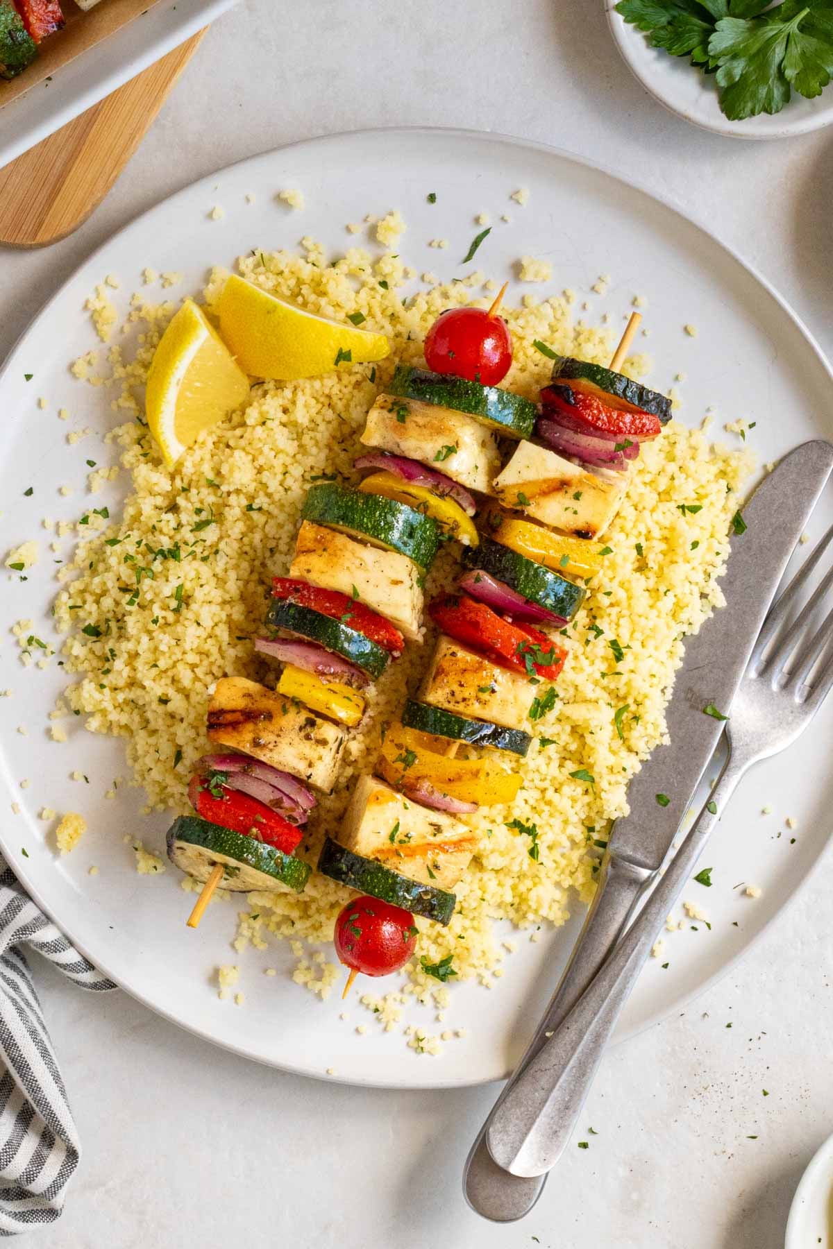 A large white plate with grilled halloumi and vegetable skewers on a bed of couscous with lemon wedges on the side and silver cutlery.
