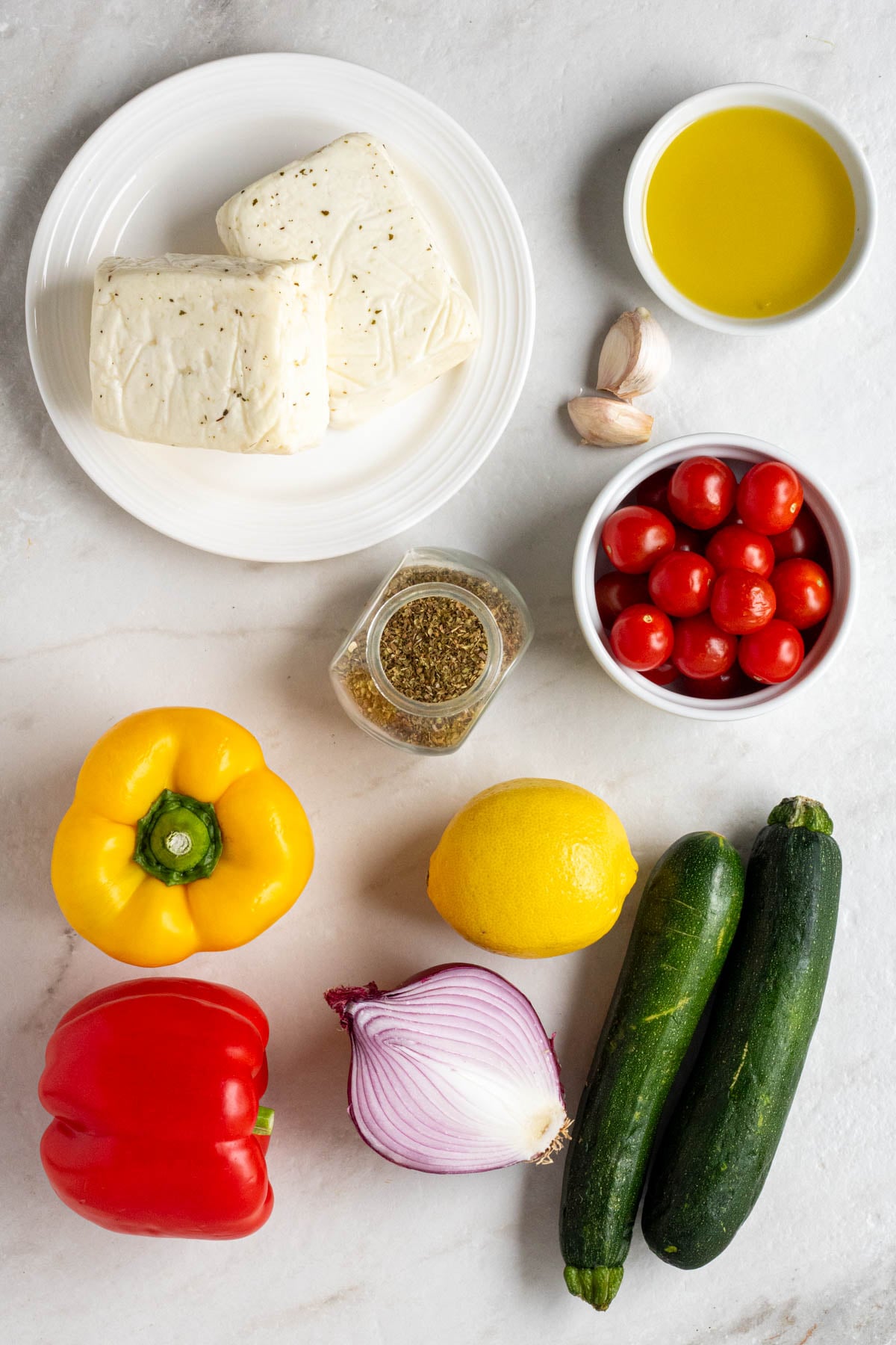 Plate of halloumi cheese, bowl of olive oil, bowl of cherry tomatoes, jar or oregano, with garlic cloves, bell peppers, red onion, lemon, and zucchini on a white background.