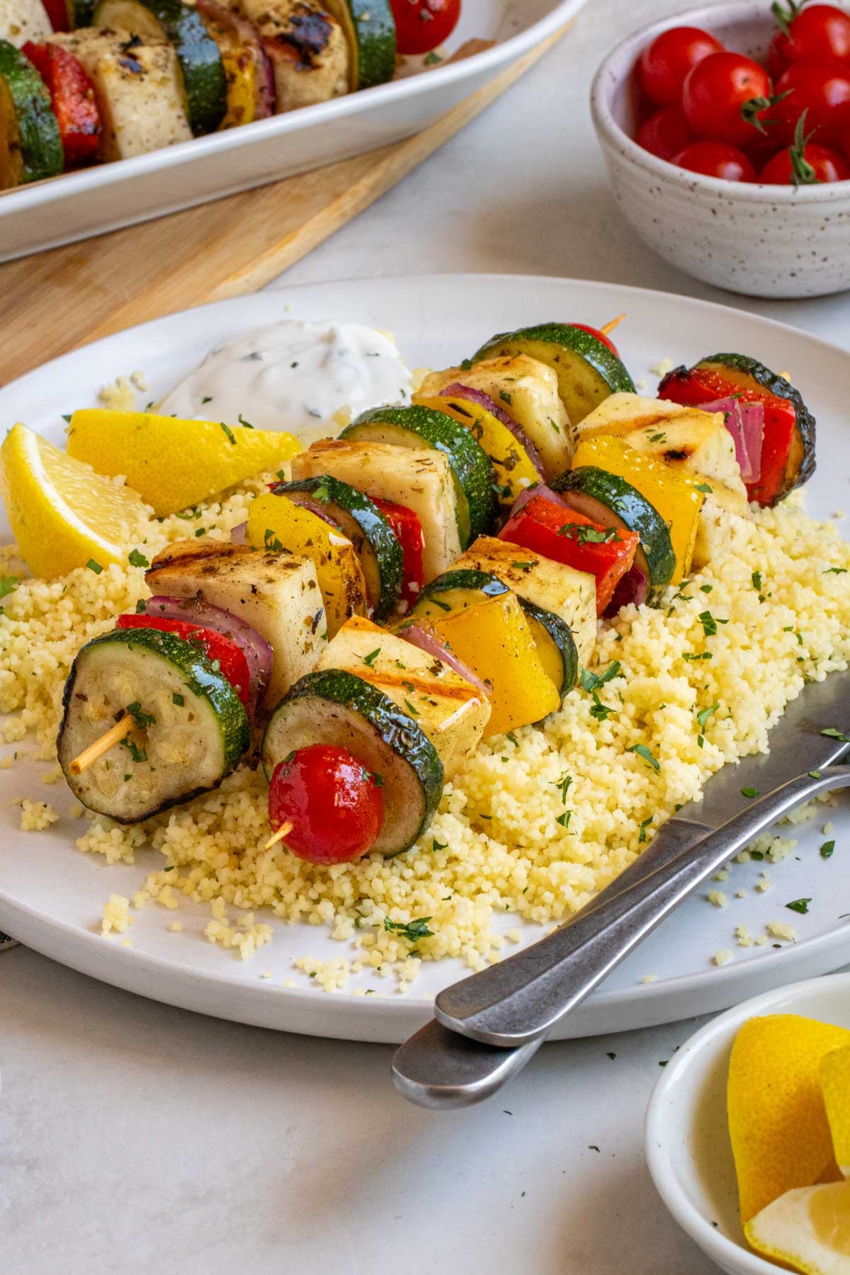 Close of up grilled veggie and halloumi skewers with couscous, tzatziki, and lemon wedges.