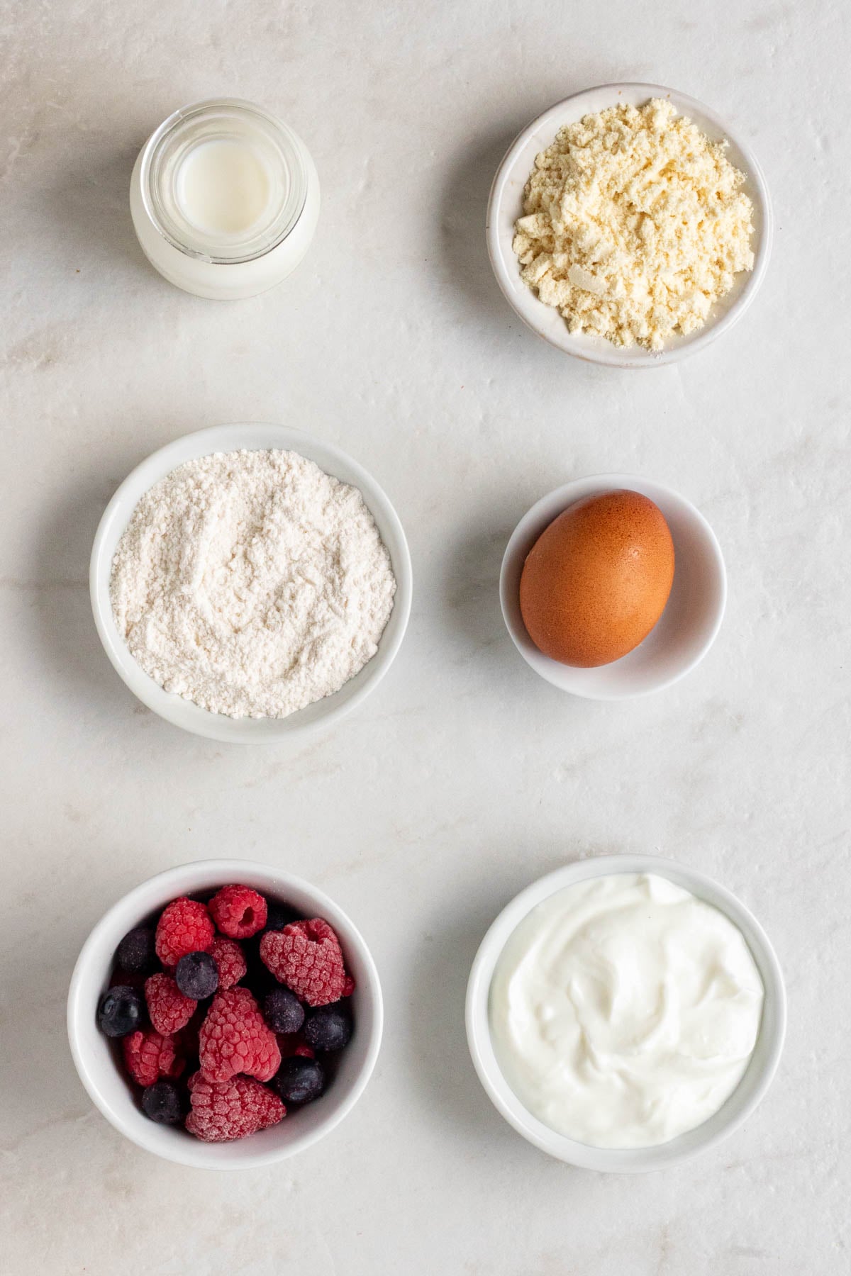 Jar of milk, bowl of protein powder, bowl of flour, egg, bowl of frozen berries, and bowl of yogurt on a white background.