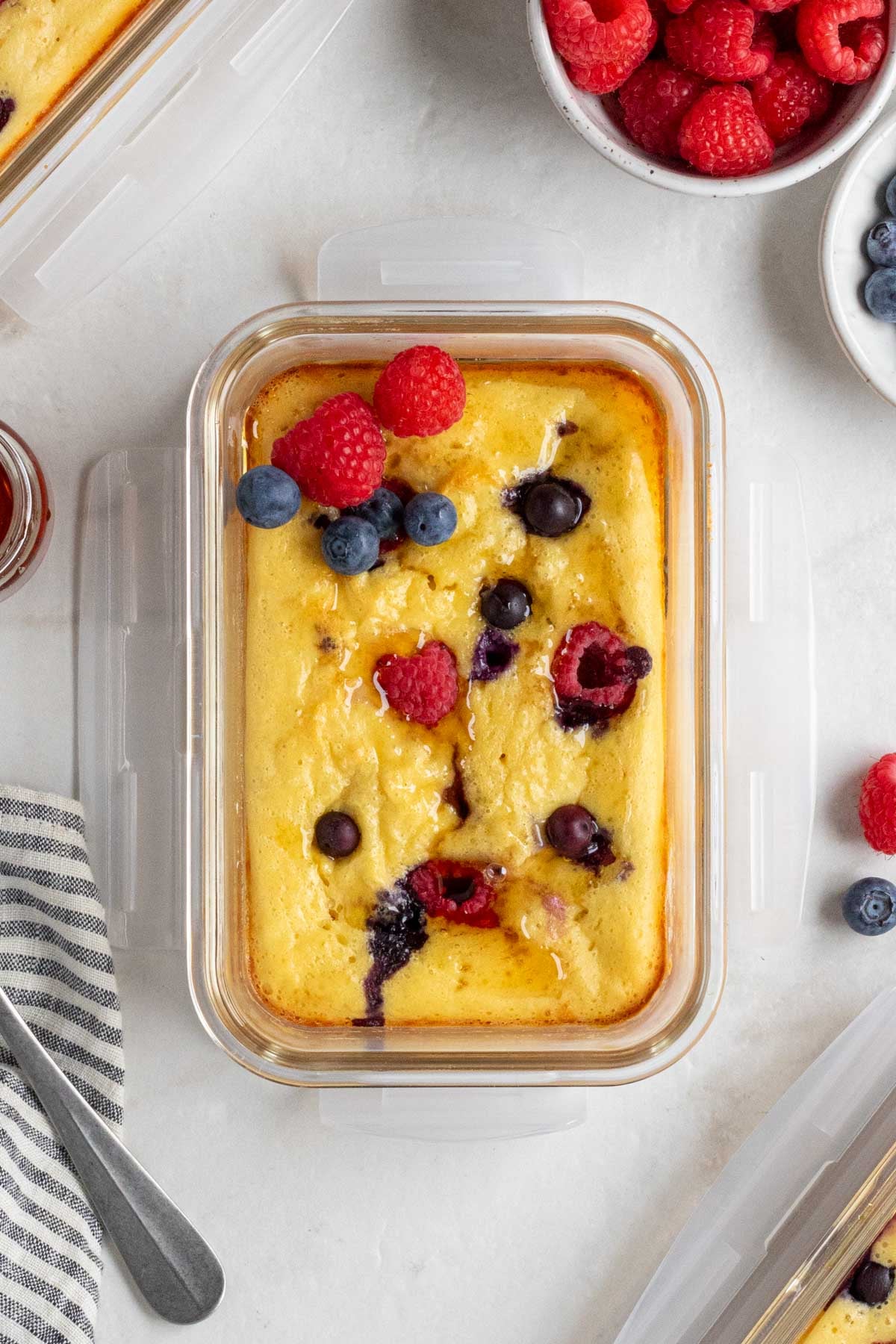 Protein pancake bowl with berries and maple syrup on top with a blue and white striped tea towel and silver spoon on the side.