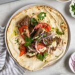 Plate with a sheet pan beef kebab pita with garlic tahini sauce, sliced tomatoes, red onion, and parsley on top on a grey and white tea towel.