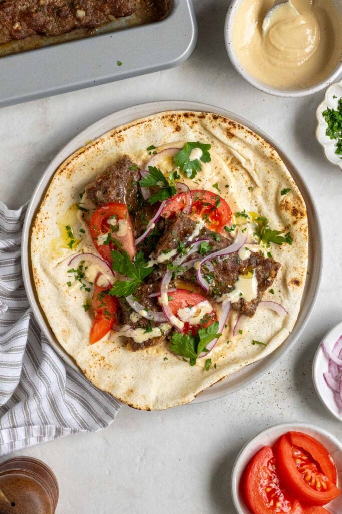 Plate with a sheet pan beef kebab pita with garlic tahini sauce, sliced tomatoes, red onion, and parsley on top on a grey and white tea towel.