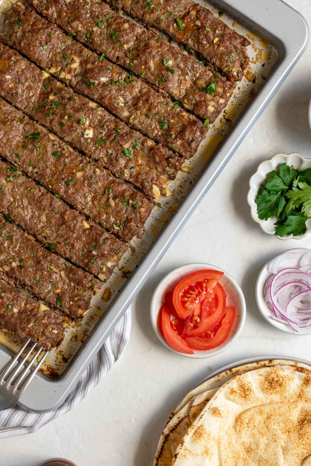 Baking sheet with slices of sheet pan beef kebabs with bowls of parsley, tomato slices, and sliced red onion on the side with a plate of pitas.