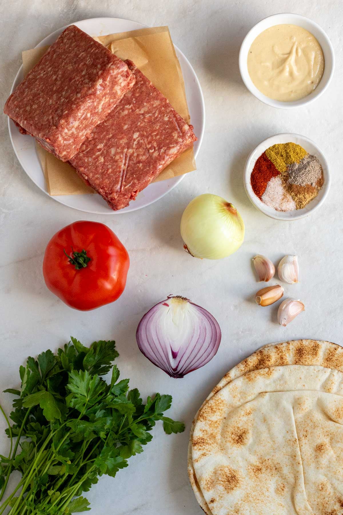 Plate of lean ground beef, bowl of garlic tahini sauce, bowl of spices, tomato, red onion, garlic, parsley, and pitas on a white background.