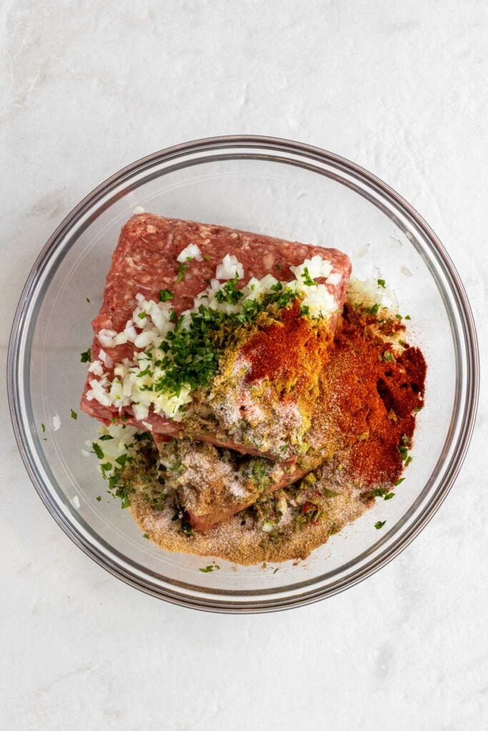 Glass bowl with lean ground beef, minced onion, minced garlic, chopped parsley, and spices on a white background.