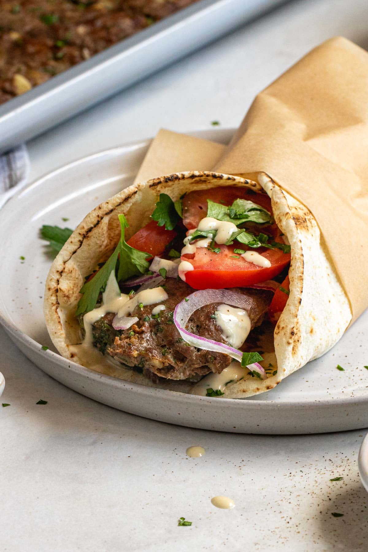 Beef kebab pita wrap on a plate with tomatoes, red onion, parsley, and tahini sauce.