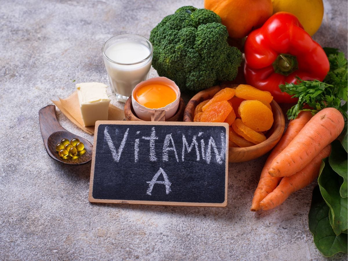 Chalkboard with vitamin A written on it with tofu, milk, egg yolk, broccoli, apricots, carrots, bell pepper, pumpkin, squash, and parsley on a grey background.