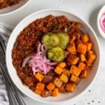 Two sloppy joe bowls with a bowl of steamed broccoli and pickled red onions on the side and a grey and white striped tea towel with a silver fork and spoon.