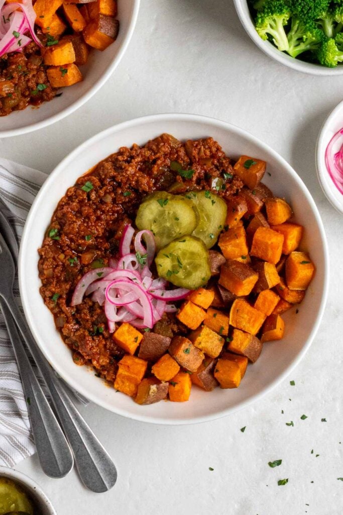 Two sloppy joe bowls with a bowl of steamed broccoli and pickled red onions on the side and a grey and white striped tea towel with a silver fork and spoon.