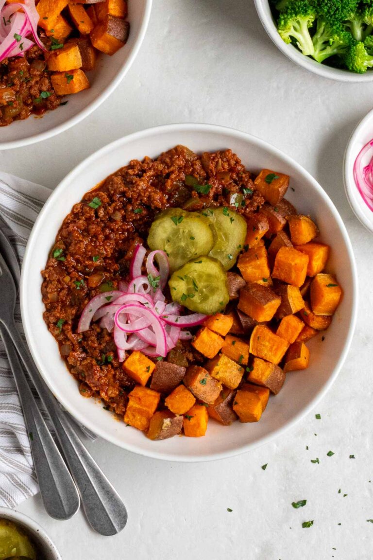 Two sloppy joe bowls with a bowl of steamed broccoli and pickled red onions on the side and a grey and white striped tea towel with a silver fork and spoon.