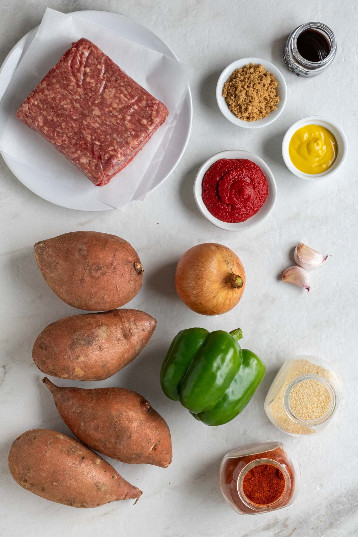 Plate of lean ground beef, jar of soy sauce, bowl of brown sugar, bowl of mustard, bowl of tomato paste, onion, garlic cloves, green bell pepper, sweet potatoes, and a jar of paprika and garlic powder on a white background.