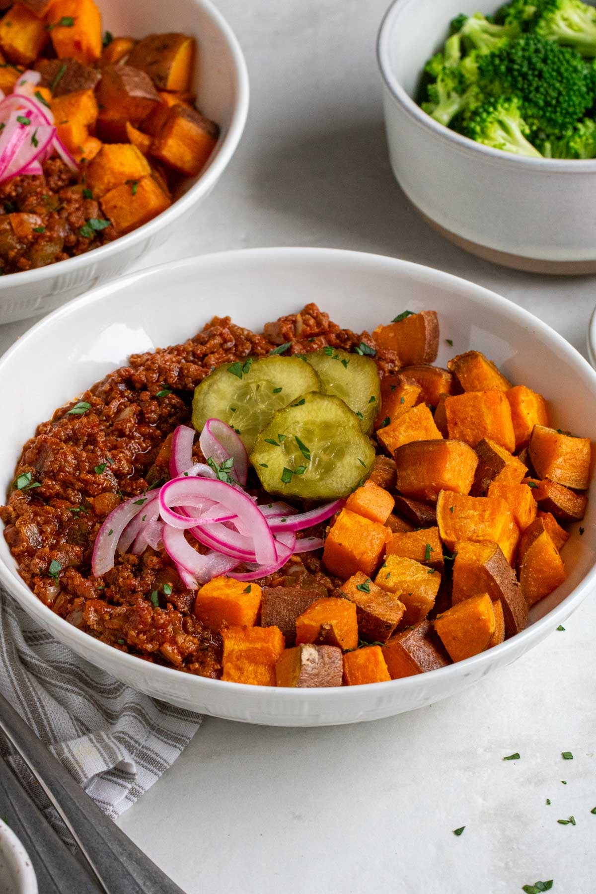 Close up of a bowl of sloppy joe meat with roasted sweet potatoes, pickles, pickled red onions, and chopped parsley on top.