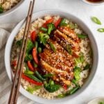Two teriyaki chicken and rice bowls on a beige striped tea towel with dark wood chopsticks and a small bowl of teriyaki sauce and bowl of sliced green onion on the side.