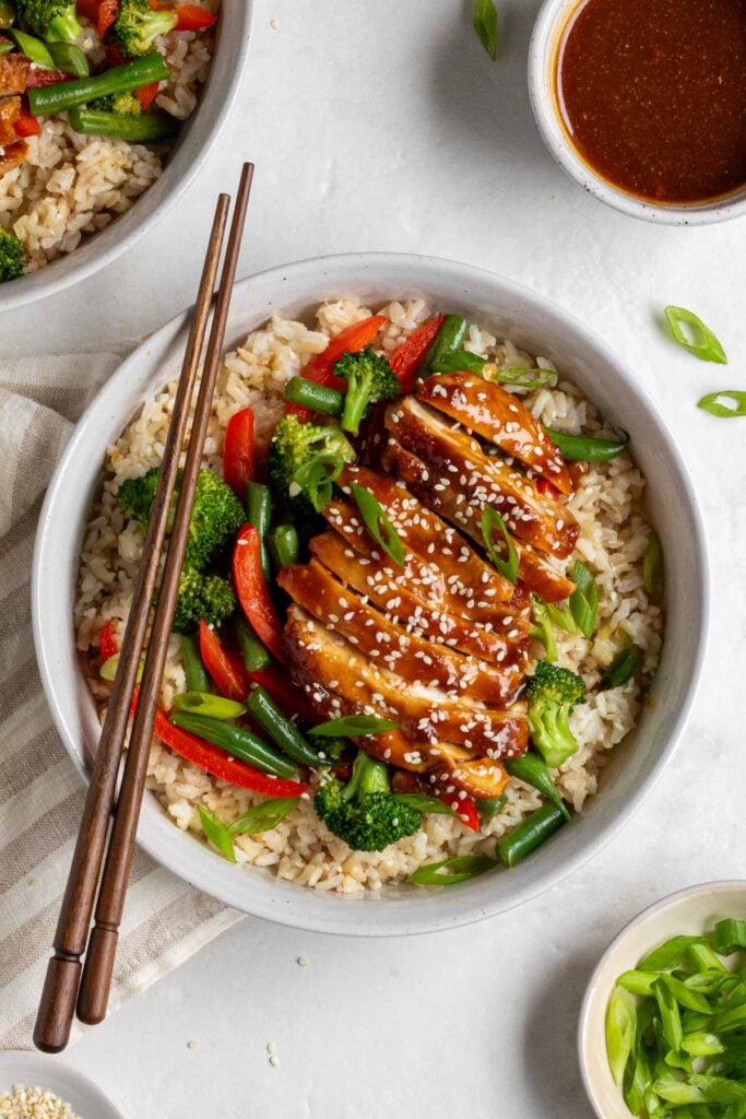 Two teriyaki chicken and rice bowls on a beige striped tea towel with dark wood chopsticks and a small bowl of teriyaki sauce and bowl of sliced green onion on the side.