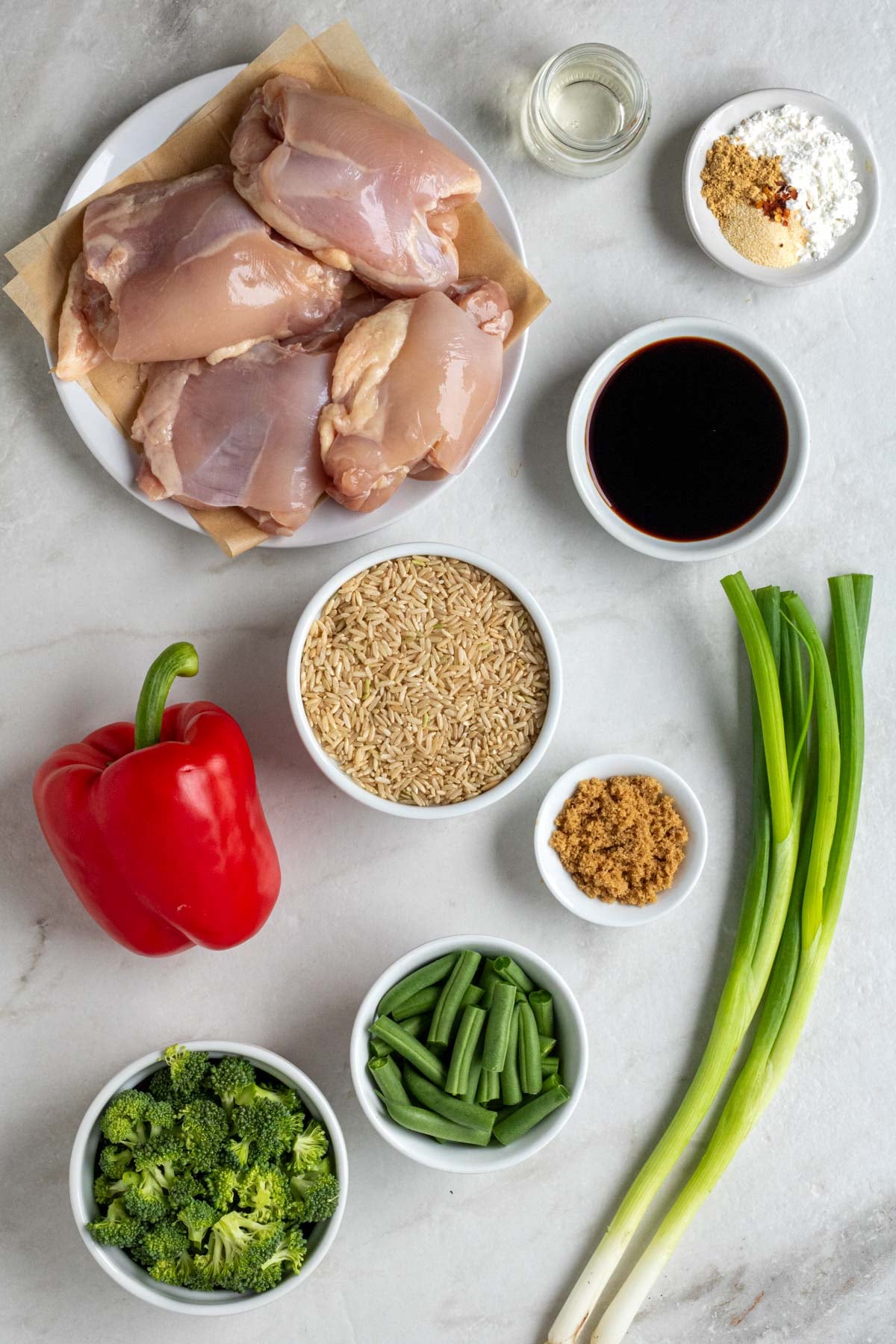 Plate of chicken thighs, jar of rice vinegar, bowl of spices and cornstarch, bowl of brown rice, bowl of brown sugar, bowl of green beans, bowl of broccoli florets, green onions, and red bell pepper on a white background.
