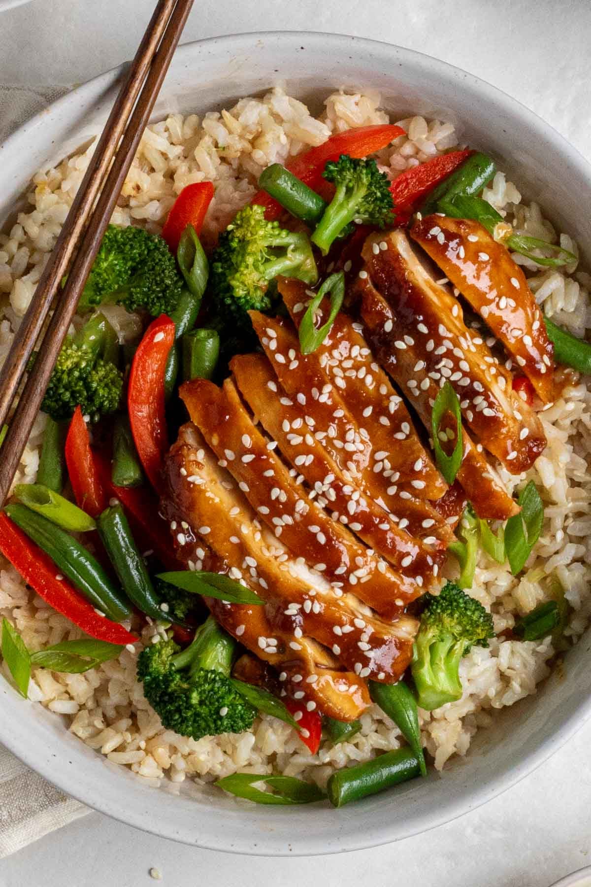 Close up of a bowl of brown rice, with stir-fried vegetables, and a cooked chicken thigh covered in teriyaki sauce with sesame seeds on top.