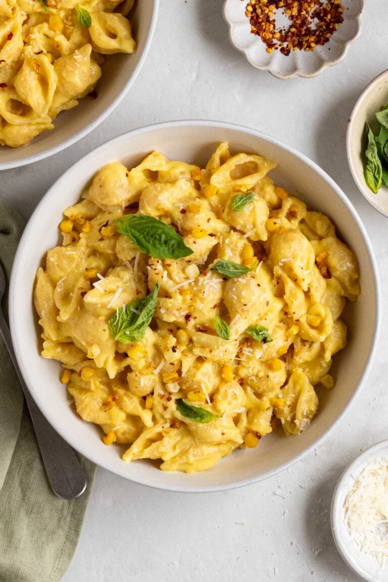 Two bowls creamy corn pasta with fresh basil on top on a light green tea towel with a fork on the side and small bowls of red pepper flakes and freshly grated parmesan cheese on the side.