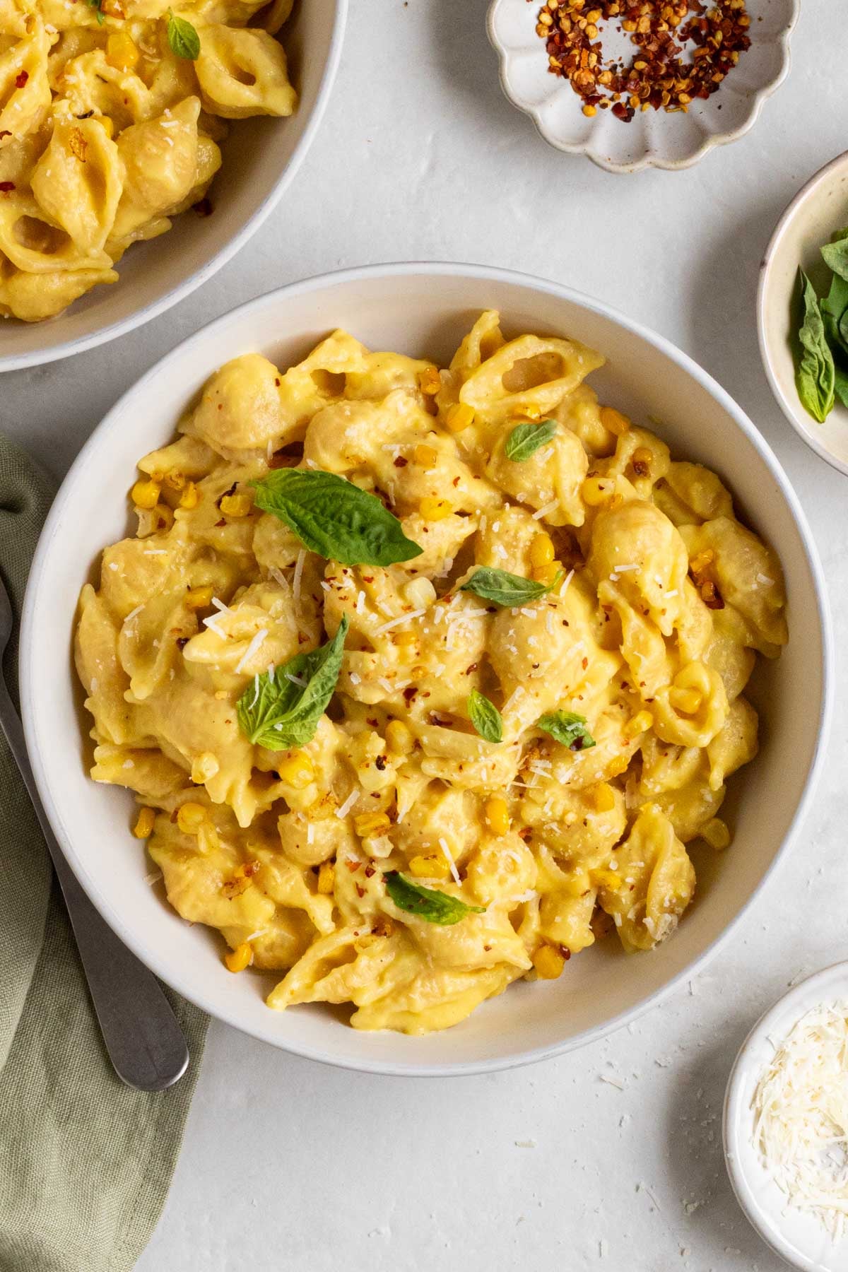 Two bowls creamy corn pasta with fresh basil on top on a light green tea towel with a fork on the side and small bowls of red pepper flakes and freshly grated parmesan cheese on the side.