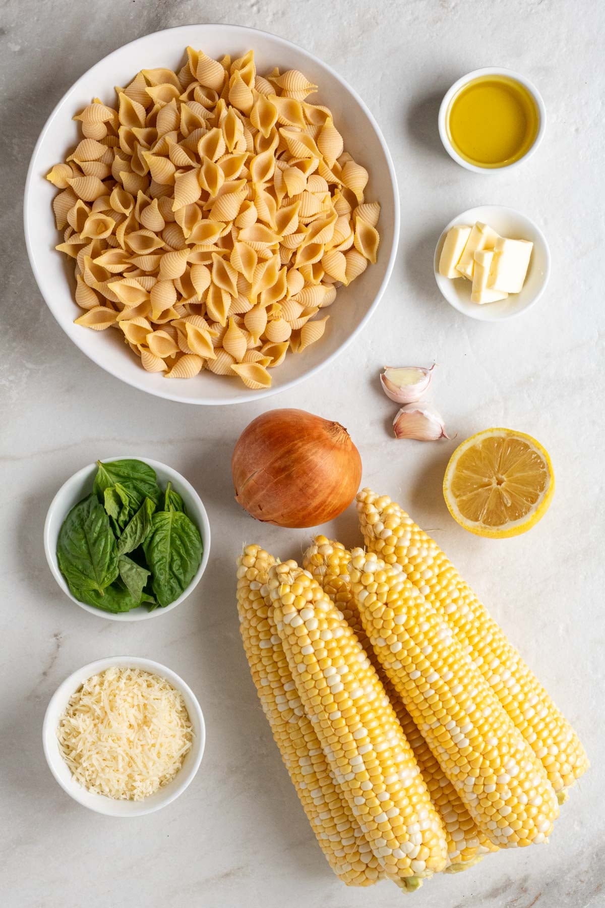Large bowl of uncooked shell pasta, small bowls of olive oil and butter, garlic cloves, fresh ears of corn, bowl of fresh basil, and a bowl of freshly grated parmesan cheese on a white background.