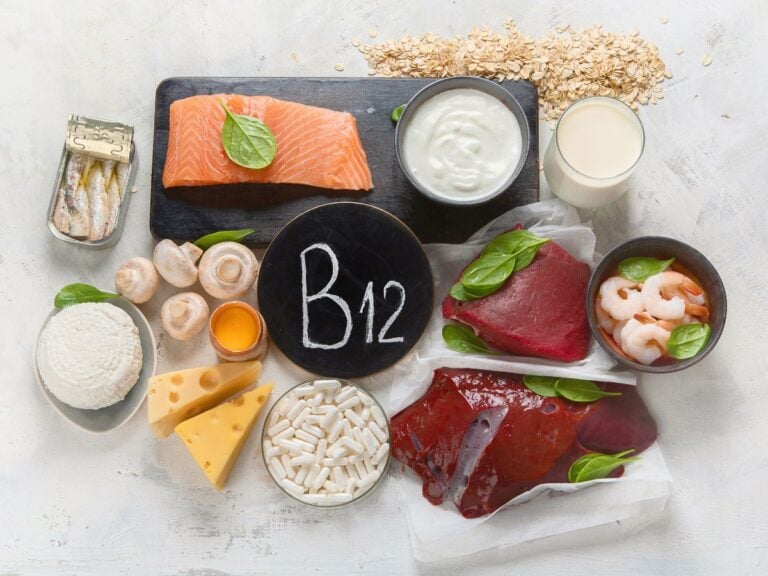 Chalkboard with B12 written on it surrounded by sardines, salmon, yogurt, oats, soy milk, shrimp , tuna, beef liver, pills, Swiss cheese, egg, mushrooms, ricotta cheese.