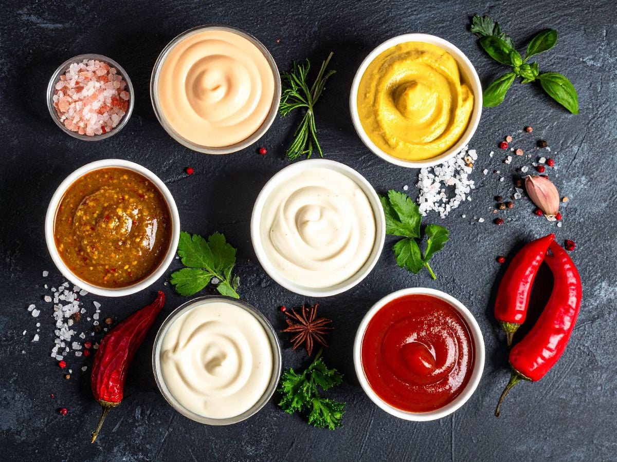 Small bowls of spicy mayo, mustard, honey mustard, Greek yogurt, mayonnaise, and ketchup on a dark background.