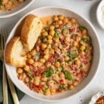 Two bowls of marry me chickpeas with crusty bread, fresh basil, and freshly grated parmesan cheese on top with a gold fork and spoon on a dark green tea towel and a small bowl of red pepper flakes on the side.
