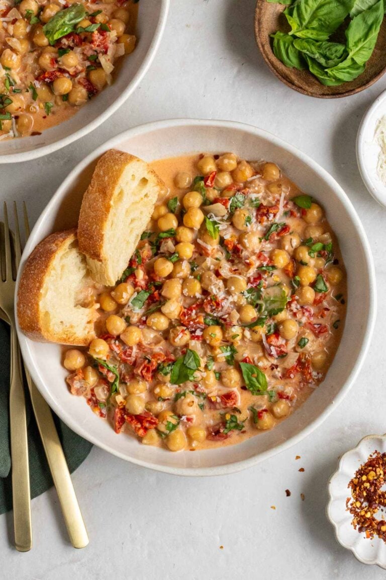 Two bowls of marry me chickpeas with crusty bread, fresh basil, and freshly grated parmesan cheese on top with a gold fork and spoon on a dark green tea towel and a small bowl of red pepper flakes on the side.