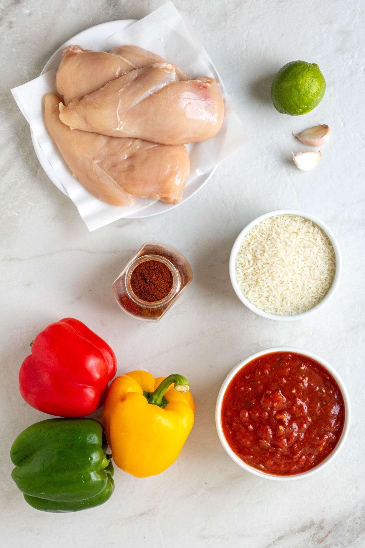 Plate of chicken breasts, lime, garlic cloves, bowl of white rice, jar of chili powder, bell peppers, and bowl of salsa on a white background.